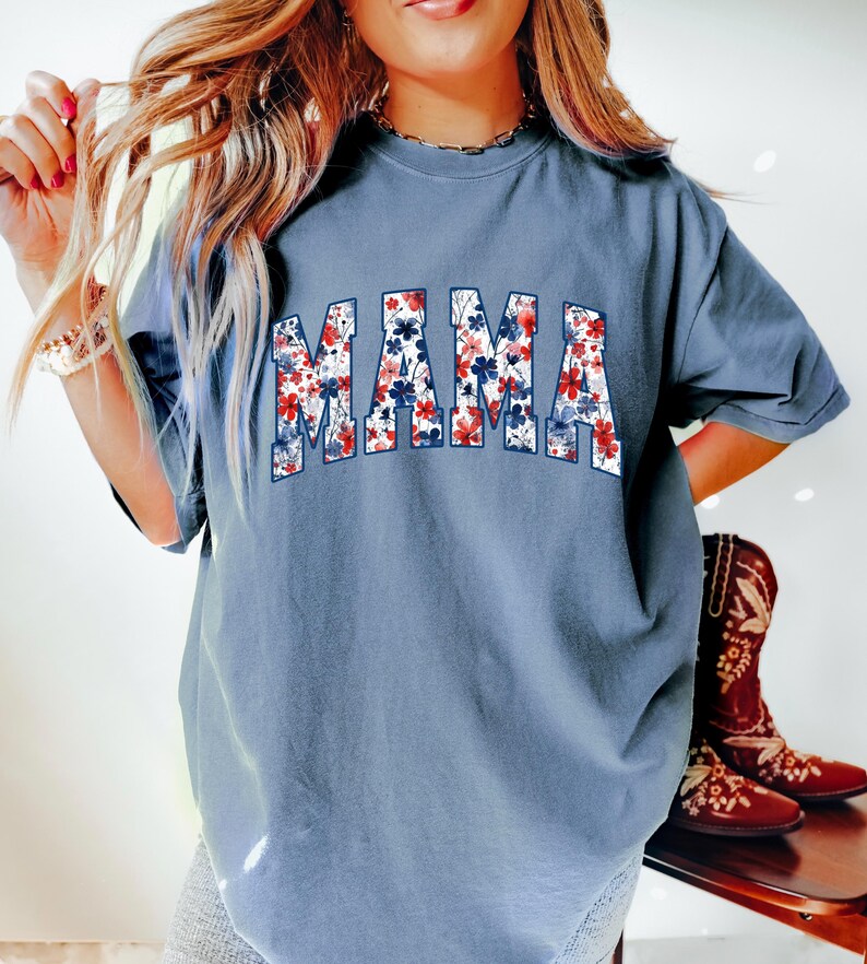 May include: A dusty blue t-shirt with the word "MAMA" in a floral design. The letters are filled with red, white, and blue flowers. The shirt has short sleeves and a relaxed fit. The image also shows a pair of brown cowboy boots.
