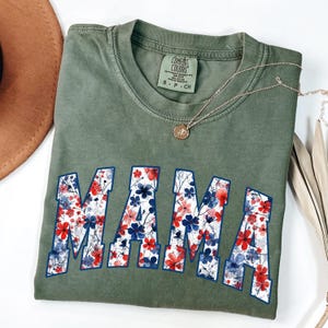May include: Olive green t-shirt with the word "MAMA" in large, floral-filled letters. The flowers are red and blue with a white background. A gold necklace with a pendant is draped across the shirt.