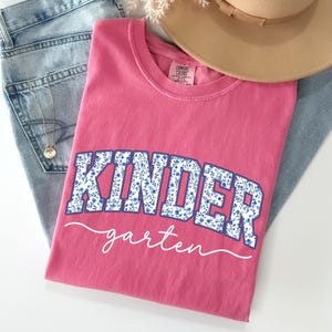 May include: A pink t-shirt with the word "KINDER" in blue floral print letters and "garten" in white script. The shirt is folded on top of a pair of blue jeans and a tan hat.