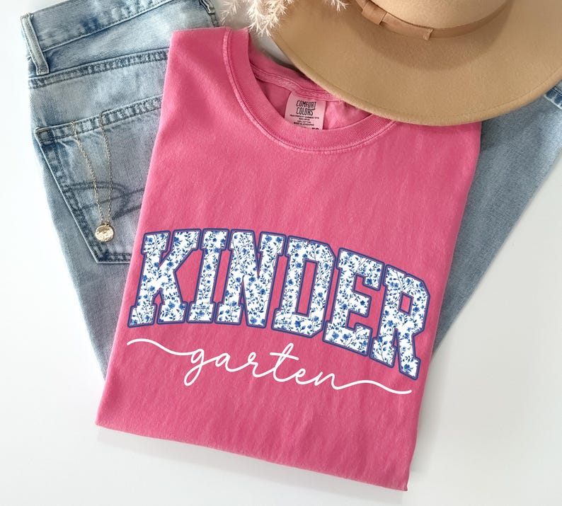 May include: A pink t-shirt with the word "KINDER" in blue floral print letters and "garten" in white script. The shirt is folded on top of a pair of blue jeans and a tan hat.