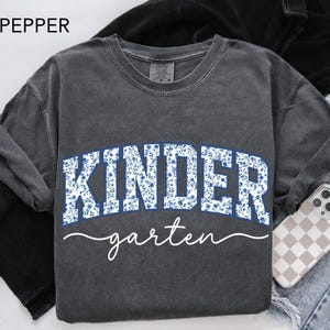 May include: A dark gray sweatshirt with the word "KINDER" in white block letters filled with a blue floral pattern. The word "garten" is written in a cursive font below. The word "PEPPER" is in the top left corner.