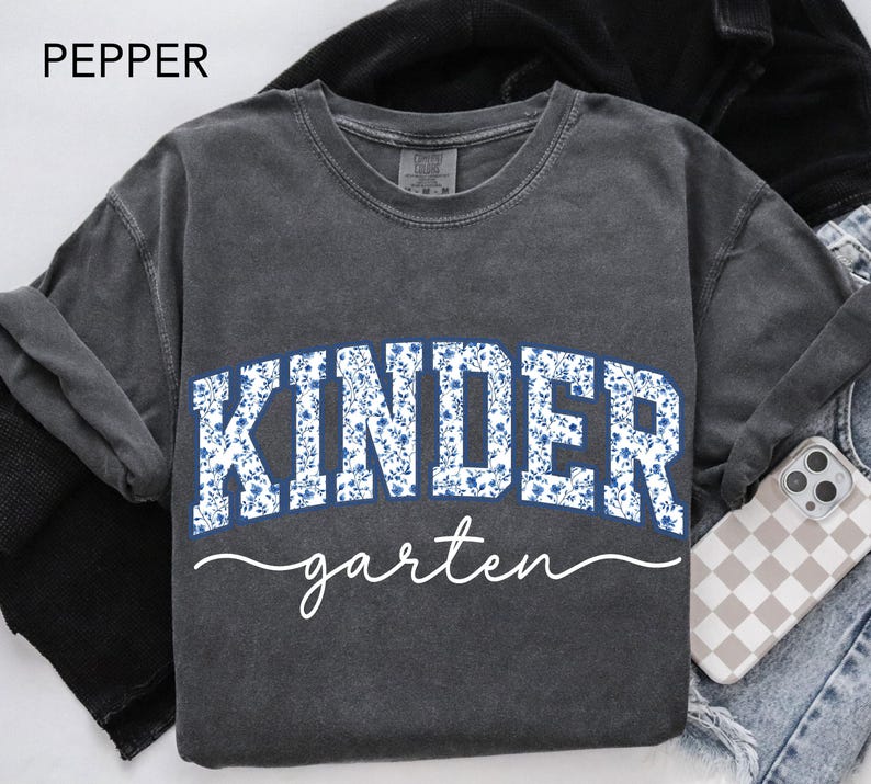 May include: A dark gray sweatshirt with the word "KINDER" in white block letters filled with a blue floral pattern. The word "garten" is written in a cursive font below. The word "PEPPER" is in the top left corner.