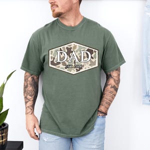 May include: Olive green t-shirt with a camouflage design in a hexagon shape, featuring the word "DAD" and "EST. 2025" in a serif font. The shirt is a casual style.