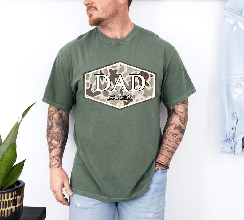 May include: Olive green t-shirt with a camouflage design in a hexagon shape, featuring the word "DAD" and "EST. 2025" in a serif font. The shirt is a casual style.