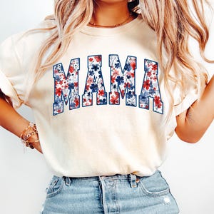 May include: Cream-colored t-shirt with the word "MAMA" in large, block letters. The letters are filled with a floral pattern in red, white, and blue, creating a patriotic design. The shirt has short sleeves and a relaxed fit.