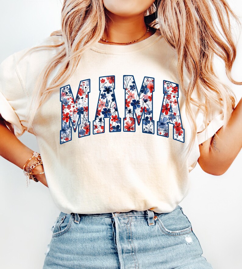 May include: Cream-colored t-shirt with the word "MAMA" in large, block letters. The letters are filled with a floral pattern in red, white, and blue, creating a patriotic design. The shirt has short sleeves and a relaxed fit.