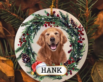 May include: A round, white Christmas ornament with a golden retriever dog inside a green wreath. The wreath is decorated with red berries, pine cones, and a Santa hat. The ornament has the name "HANK" written on it.