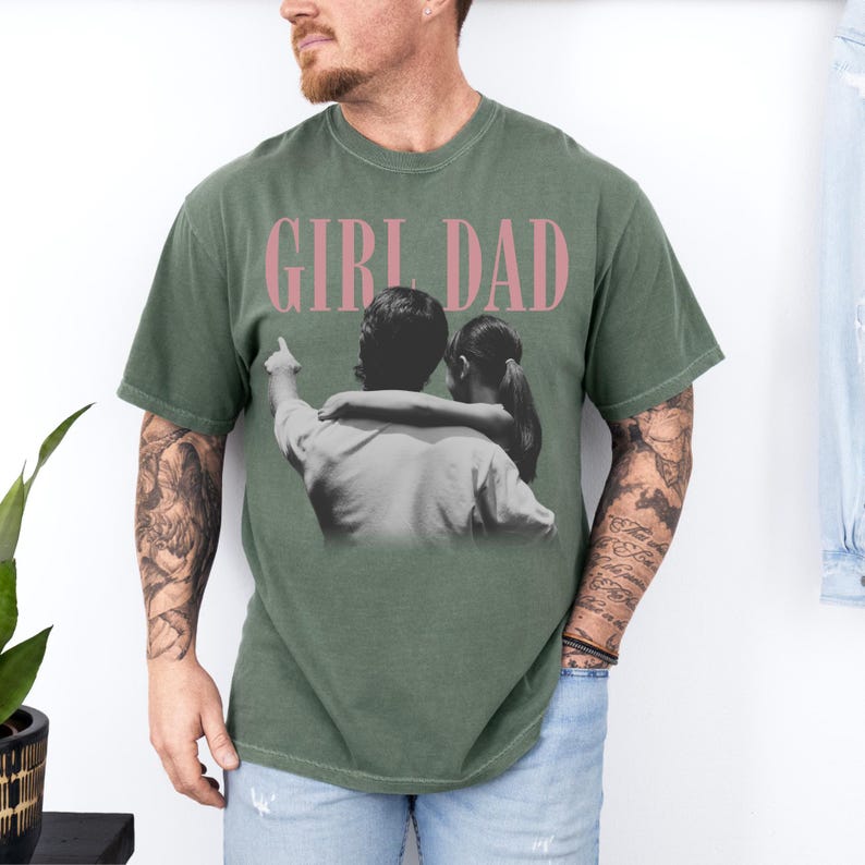 K&ouml;nnte beinhalten: Olivgr&uuml;nes T-Shirt mit dem rosa Schriftzug "GIRL DAD". Das Shirt zeigt ein Schwarz-Wei&szlig;-Bild eines Vaters und seiner Tochter. Der Vater zeigt nach oben, w&auml;hrend die Tochter ihn umarmt.