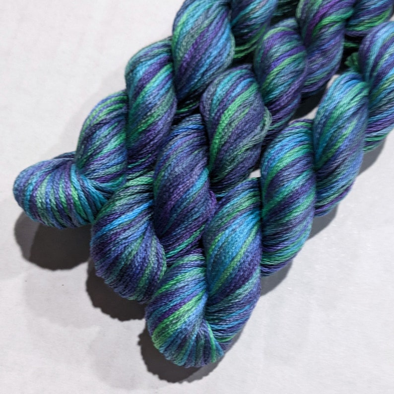 May include: Two skeins of yarn in a blue, green, and purple colorway. The yarn is twisted and folded, showing the different colors.