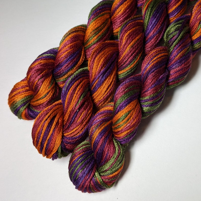 May include: Three skeins of embroidery floss in a multi-coloured pattern of deep purple, orange, and green.