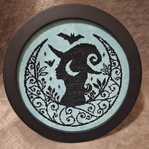 May include: A black and white cross-stitch embroidery framed in a black circular frame. The design features a silhouette of a woman with a crescent moon and a bat in the background.