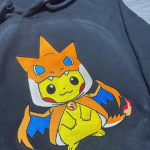 May include: Black hooded sweatshirt with a Pikachu character in a Charizard costume. The design features orange, blue, and white embroidery. The hoodie has a drawstring and a front pocket. The design is a cartoon style.