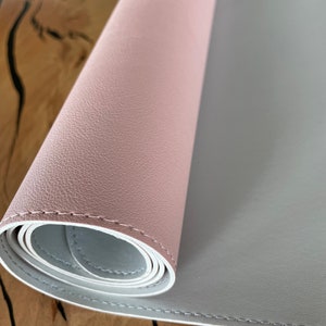 Pu&ograve; includere: Un tappetino da scrivania bicolore arrotolato. Lo strato superiore &egrave; rosa tenue e lo strato inferiore &egrave; grigio chiaro. I bordi sono cuciti con filo bianco. Il tappetino &egrave; su una superficie di legno.