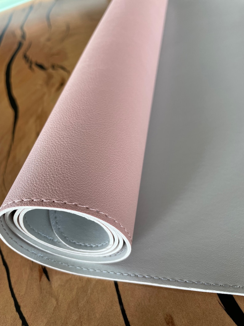 Pu&ograve; includere: Un tappetino da scrivania bicolore arrotolato. Lo strato superiore &egrave; rosa tenue e lo strato inferiore &egrave; grigio chiaro. I bordi sono cuciti con filo bianco. Il tappetino &egrave; su una superficie di legno.