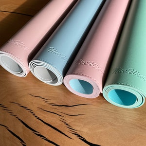 Pu&ograve; includere: Quattro tovagliette arrotolate in colori pastello: rosa, azzurro, rosa e verde menta. Ogni tappetino ha un lato inferiore bianco e reca la scritta "Nice Wear". Le tovagliette sono disposte su una superficie di legno.