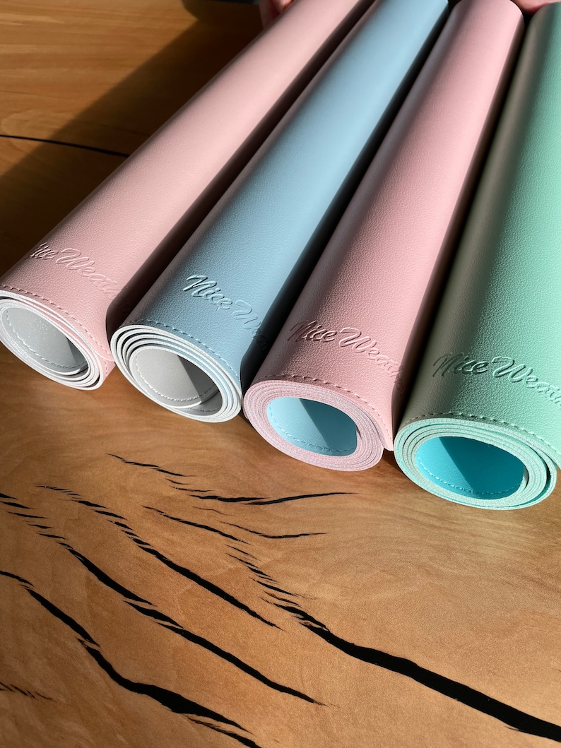 Pu&ograve; includere: Quattro tovagliette arrotolate in colori pastello: rosa, azzurro, rosa e verde menta. Ogni tappetino ha un lato inferiore bianco e reca la scritta "Nice Wear". Le tovagliette sono disposte su una superficie di legno.