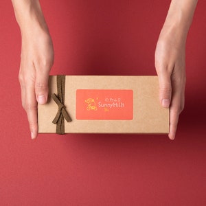 May include: A rectangular cardboard gift box with a brown ribbon tied in a bow. The box has a red sticker with the text "Sunny Hills" and a cartoon image. The box is held by two hands against a red background.