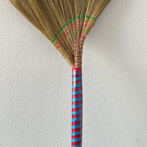 May include: A handmade broom with a fan-shaped head made of natural fibers. The handle is wrapped with alternating red and blue bands. Green and pink rubber bands secure the broom head.