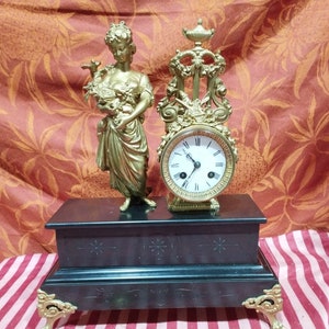 Peut inclure: Une horloge de cheminée dorée avec une base en marbre noir et une figurine de femme tenant un oiseau. Le cadran de l'horloge a des chiffres romains et un cadre doré.