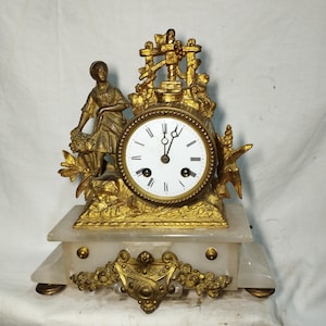 May include: Antique gold-toned mantel clock with a white clock face and black Roman numerals. The clock features a decorative woman figure and ornate floral accents. The clock sits on a white marble base with gold embellishments.