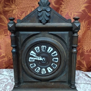 Peut inclure: Une horloge en bois brun foncé avec un cadran blanc et des chiffres romains noirs. L'horloge a un sommet en bois sculpté et des détails ornés.