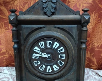 May include: A dark brown wooden clock with a white face and black Roman numeral hour markers. The clock has a carved wooden top and ornate details.