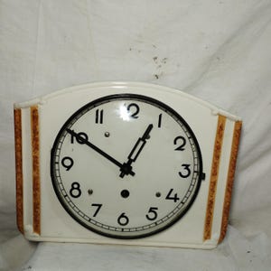 Puede incluir: Reloj rectangular color crema con esfera redonda. La esfera del reloj tiene números y manecillas negras. El marco del reloj tiene rayas verticales marrones a cada lado. El reloj mide aproximadamente 25 cm de ancho y 18 cm de alto.