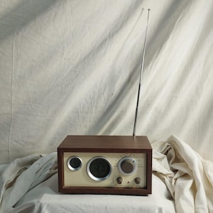 May include: A vintage-style radio with a wooden cabinet and a cream-colored face. The radio features a digital clock display, tuning dials, and a retractable antenna. The design is retro, with a focus on simplicity and functionality.