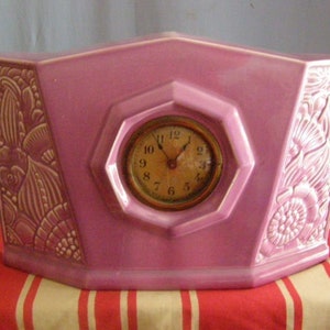 May include: A pink ceramic clock with a floral design and a white clock face with black numbers. The clock is shaped like a house and has a curved base.