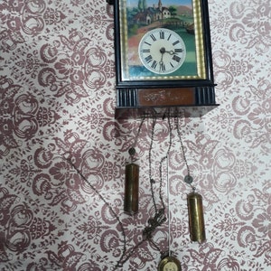 Puede incluir: Un reloj de pared de madera negra con una esfera dorada y una escena pintada de un pueblo. El reloj tiene una cadena y pesas colgando.