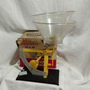 Peut inclure: Une balance de cuisine vintage en métal jaune avec un bol en plastique transparent en forme d'entonnoir. La balance présente une jauge de mesure métallique incurvée avec des graduations noires. Une boîte en carton marquée NOUVEAU PESE-PRODUITS 350 GR. MP.