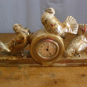 May include: A ceramic clock with three brown and gold birds perched on a base. The clock face has black Roman numeral markings and black hands.