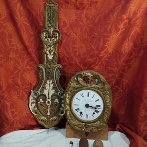 Puede incluir: Un reloj vintage con marco dorado y detalles ornamentados. La esfera del reloj es blanca con números romanos negros y una aguja negra. El reloj está colgado en una pared roja con un patrón floral.