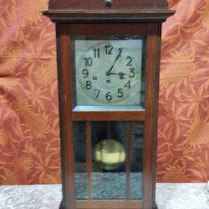 May include: A vintage wooden clock with a white face and black hands. The clock has a glass front and a pendulum inside. The clock is in a dark wood frame with a decorative top.