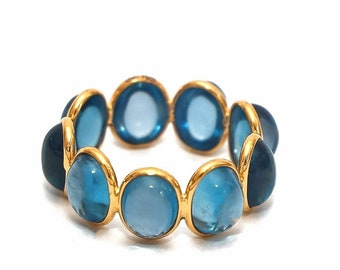 Puede incluir: Un anillo de oro con una serie de piedras preciosas ovaladas de color azul claro. Las piedras están engastadas en biseles individuales de oro, creando una banda continua de color. El anillo se muestra sobre un fondo blanco.