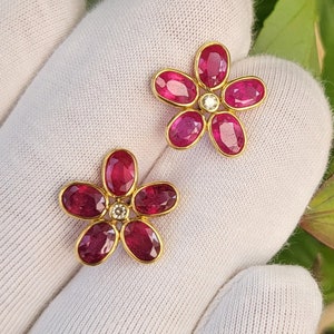 Puede incluir: Un par de pendientes de flor de rubí y diamante. Cada pendiente presenta un diseño de flor de cinco pétalos con un pequeño diamante en el centro. Los pétalos están hechos de rubíes de forma ovalada.