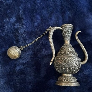 May include: A silver ornate teapot with a decorative handle and a matching lid attached by a chain. The teapot is on a blue fabric background.