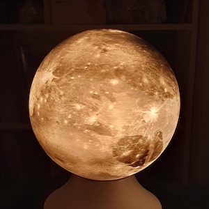 May include: A white moon lamp with a detailed surface texture, resembling the moon's surface. The lamp is on a white base and has a cord.