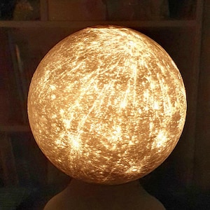 Könnte beinhalten: Eine runde, weiße Lampe mit einer strukturierten Oberfläche, die an den Mond erinnert. Die Lampe ist eingeschaltet und strahlt ein warmes, goldenes Licht aus.