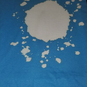 May include: A blue t-shirt with a large white bleach stain in the centre. The stain has a circular shape with smaller white splotches around it.