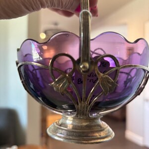 May include: A purple glass bowl with a scalloped rim, held within a silver-toned metal frame featuring a pedestal base and a vertical handle. The metal frame is decorated with floral motifs at the sides where the handle attaches to the bowl.