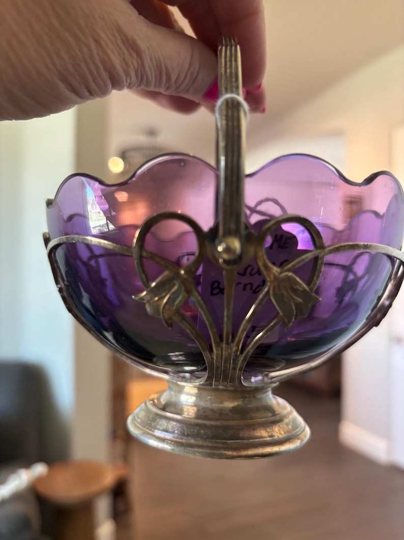 May include: A purple glass bowl with a scalloped rim, held within a silver-toned metal frame featuring a pedestal base and a vertical handle. The metal frame is decorated with floral motifs at the sides where the handle attaches to the bowl.