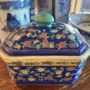 May include: A blue ceramic hexagonal trinket box with a lid. The box and lid are decorated with a floral pattern in pink, yellow, and green. The lid has a green knob. The box has a yellow border.