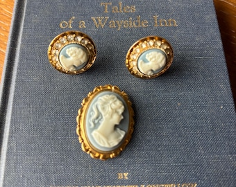 May include: A set of vintage cameo jewelry, including a brooch and two earrings, resting on a blue book. The jewelry features a gold-toned setting with a light blue and white cameo design. The book's title is "Tales of a Wayside Inn."