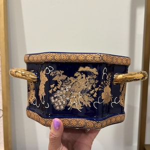 May include: A hexagonal ceramic planter featuring a deep navy blue glaze with intricate gold-toned illustrations of a peacock and floral motifs. The vessel includes two gold-toned side handles and a geometric patterned border along the top and base.