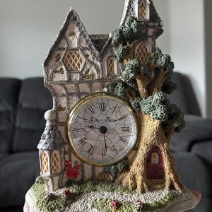 May include: A decorative clock shaped like a whimsical cottage with a tree. The clock face reads "Father Time". The cottage is beige and gray with a gold-colored clock face. The tree has a small red door.