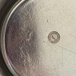 May include: A circular silver-toned metal tray with a polished, reflective surface. In the center, a small circular stamp is visible, featuring the text "ART. KRUPP BERNDORF" surrounding a central graphic of a standing bear figure.