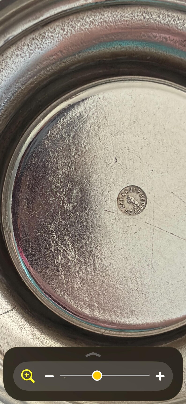 May include: A circular silver-toned metal tray with a polished, reflective surface. In the center, a small circular stamp is visible, featuring the text "ART. KRUPP BERNDORF" surrounding a central graphic of a standing bear figure.