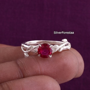 Può includere: Un anello d'argento con una gemma rotonda di colore rosso vivo. L'anello presenta un delicato design ispirato alla natura con dettagli di foglie e viti. L'anello è tenuto su uno sfondo viola.