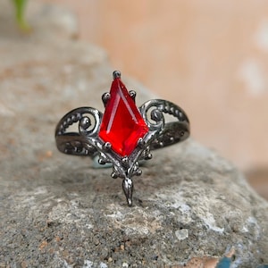 May include: A dark gunmetal-toned ring featuring a central kite-shaped red gemstone. The metal band displays intricate scrollwork and filigree detailing that extends from the base of the stone, creating a pointed, symmetrical design.